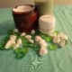 A jar of body cream, a green candle, and a small tub of Whipped Shea Butter Lavender are arranged on a teal cloth, surrounded by green and white decorative stones.