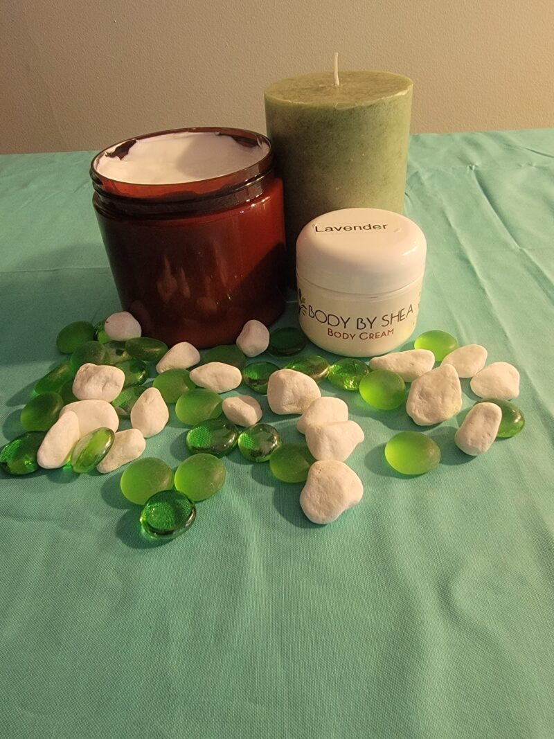 A jar of body cream, a green candle, and a small tub of Whipped Shea Butter Lavender are arranged on a teal cloth, surrounded by green and white decorative stones.
