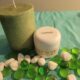 A green candle and a jar of Whipped Shea Butter Lemon are placed on a teal cloth, alongside white stones and green glass pebbles.