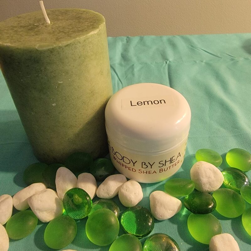 A green candle and a jar of Whipped Shea Butter Lemon are placed on a teal cloth, alongside white stones and green glass pebbles.