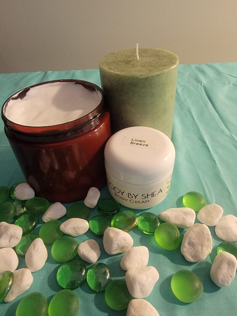 A jar of body cream, a medium-sized green candle, and a brown jar with Whipped Shea Butter Linen Breeze are placed on a blue surface adorned with green and white decorative stones.