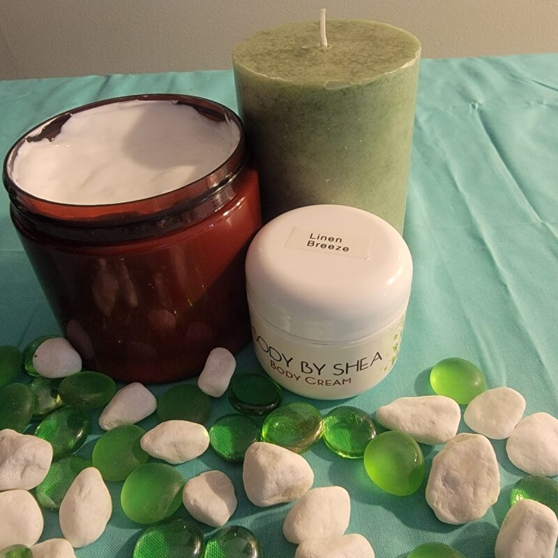 A jar of body cream, a medium-sized green candle, and a brown jar with Whipped Shea Butter Linen Breeze are placed on a blue surface adorned with green and white decorative stones.