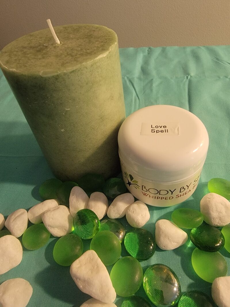 Green pillar candle and a jar labeled "Whipped Shea Butter Love Spell" on a blue surface, surrounded by white and green decorative stones.