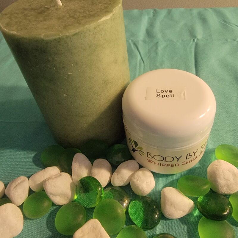 Green pillar candle and a jar labeled "Whipped Shea Butter Love Spell" on a blue surface, surrounded by white and green decorative stones.