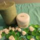 A green candle and a jar labeled "Whipped Shea Butter Mango Papaya" are placed on a turquoise fabric with green and white decorative stones scattered around.