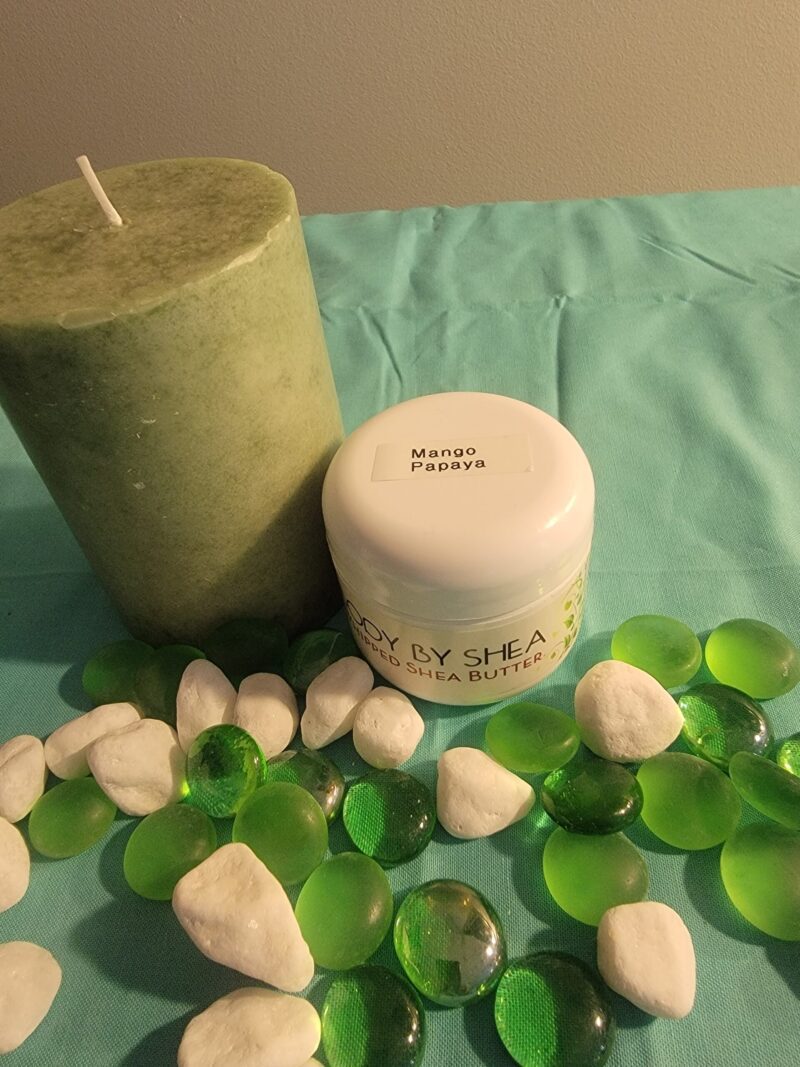 A green candle and a jar labeled "Whipped Shea Butter Mango Papaya" are placed on a turquoise fabric with green and white decorative stones scattered around.