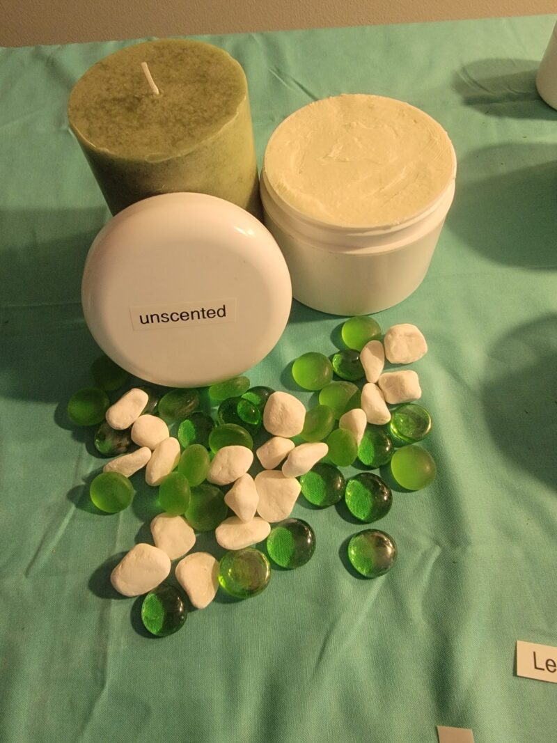 A Whipped Shea Butter Unscented and a white jar surrounded by green and white decorative stones on a teal cloth.