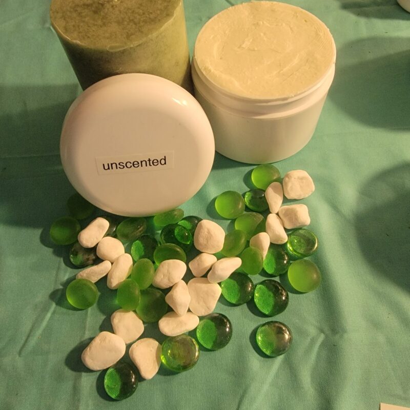 A Whipped Shea Butter Unscented and a white jar surrounded by green and white decorative stones on a teal cloth.