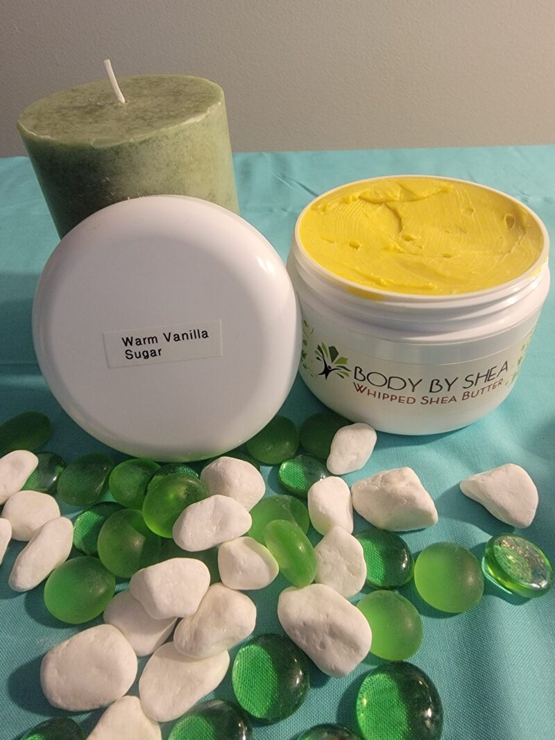 A green candle, white rocks, and green glass pebbles surround an open container of whipped shea butter labeled "Warm Vanilla Sugar." The container lid is placed next to the body butter.