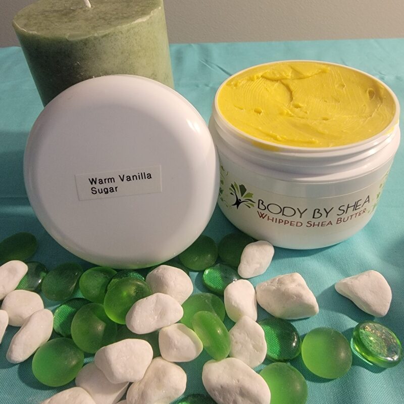 A green candle, white rocks, and green glass pebbles surround an open container of whipped shea butter labeled "Warm Vanilla Sugar." The container lid is placed next to the body butter.